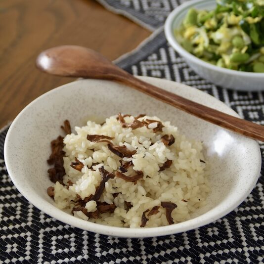 a bowl of chanterelle rice on a placemat with a wooden spoon and a side dish in the background.