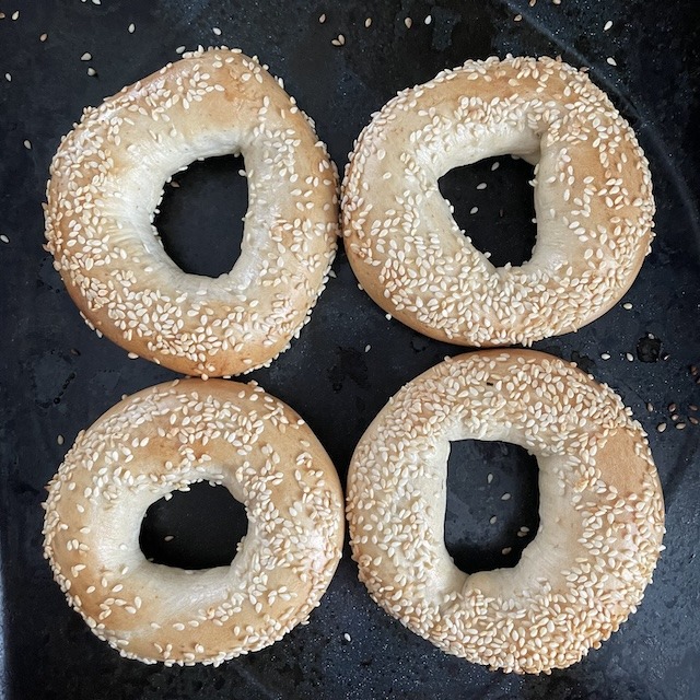 Four sesame bagels on a black baking tray.