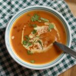 A bowl of carrot and sweet pepper soup with a metal spoon on a green plaid napkin.